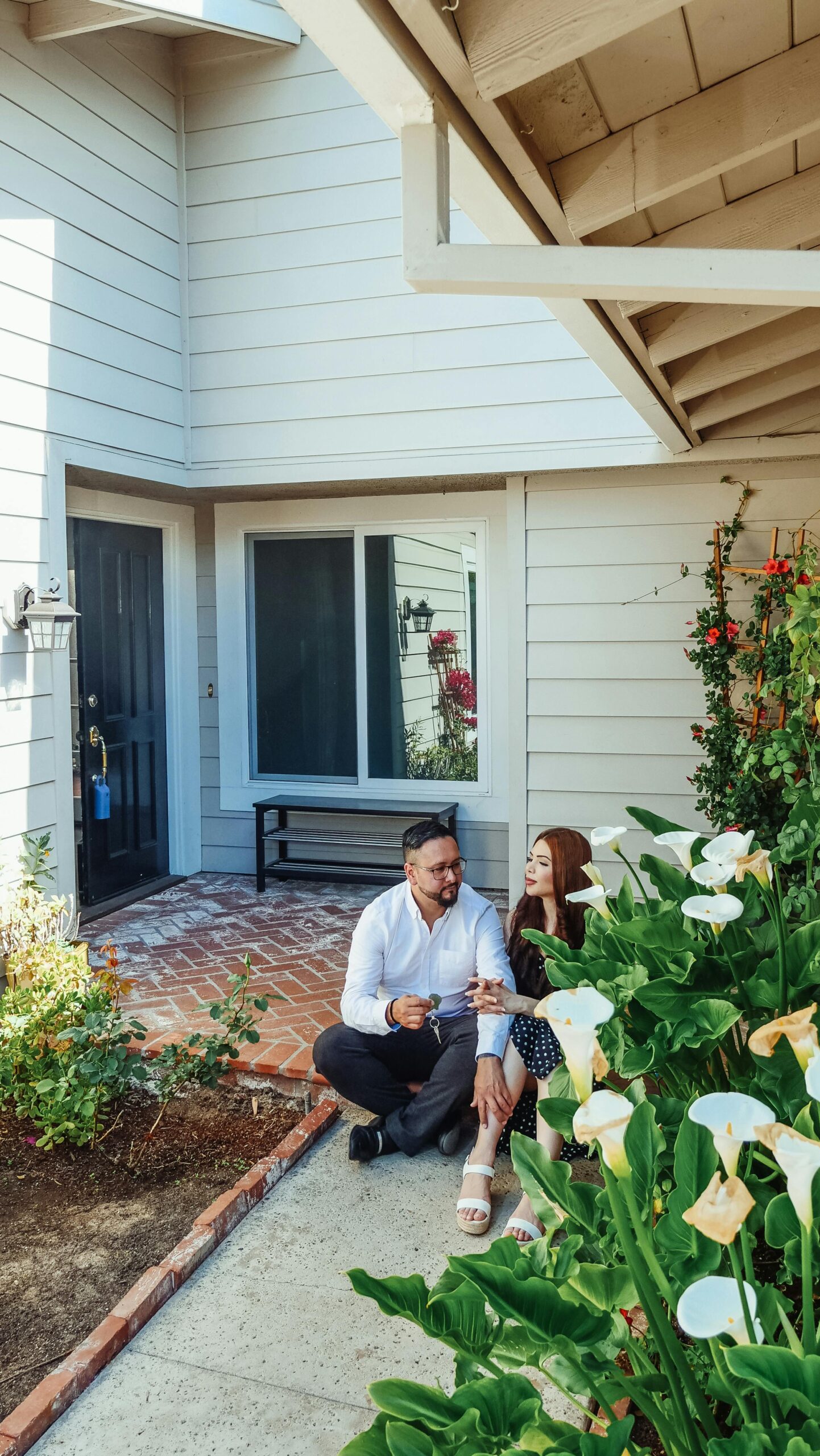 A joyful couple sits on their new home's porch surrounded by lush greenery, celebrating their purchase.